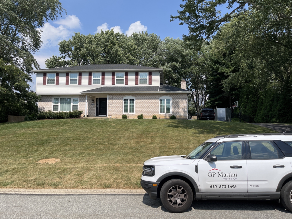A shot of a long lawn, with a split level home with a grey asphalt shingle roof and the Martini Roofing Co. truck parked in front.