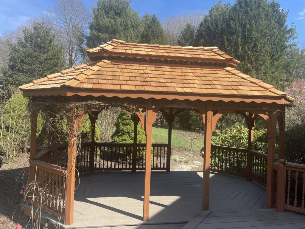 A wide, reddish-brown gazebo with plants growing around it and a cedar shake roof.